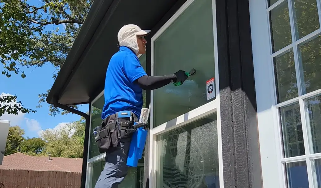 Window cleaning Bend: a cleaner in a blue shirt and cap using a squeegee on an outside window of a home