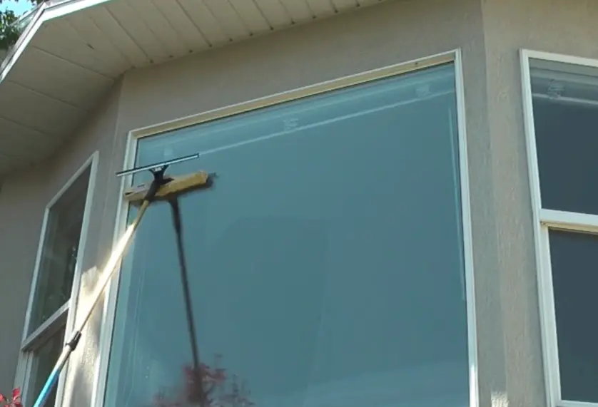 Squeegee on an extension pole cleaning a large upper window under the roof eave of a house