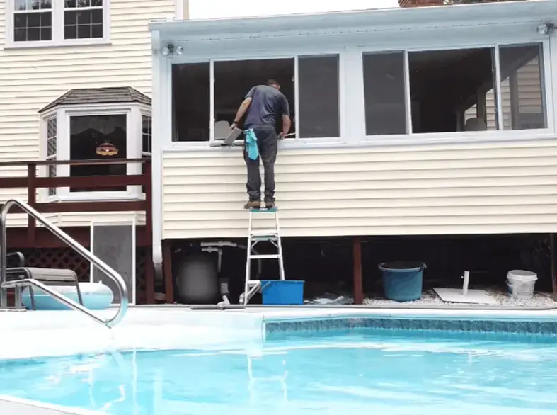 Window cleaner on a step ladder washing upper sunroom windows above a backyard pool